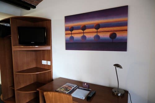 desk in hotel room with modern art hanging above and shelves with tv