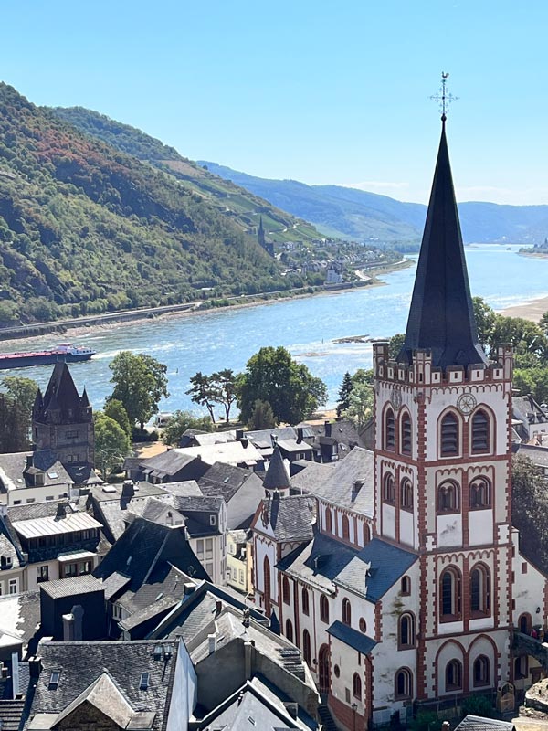 panpormaic view of church with spire and grey roofs of bacharach germany with river rhine in background