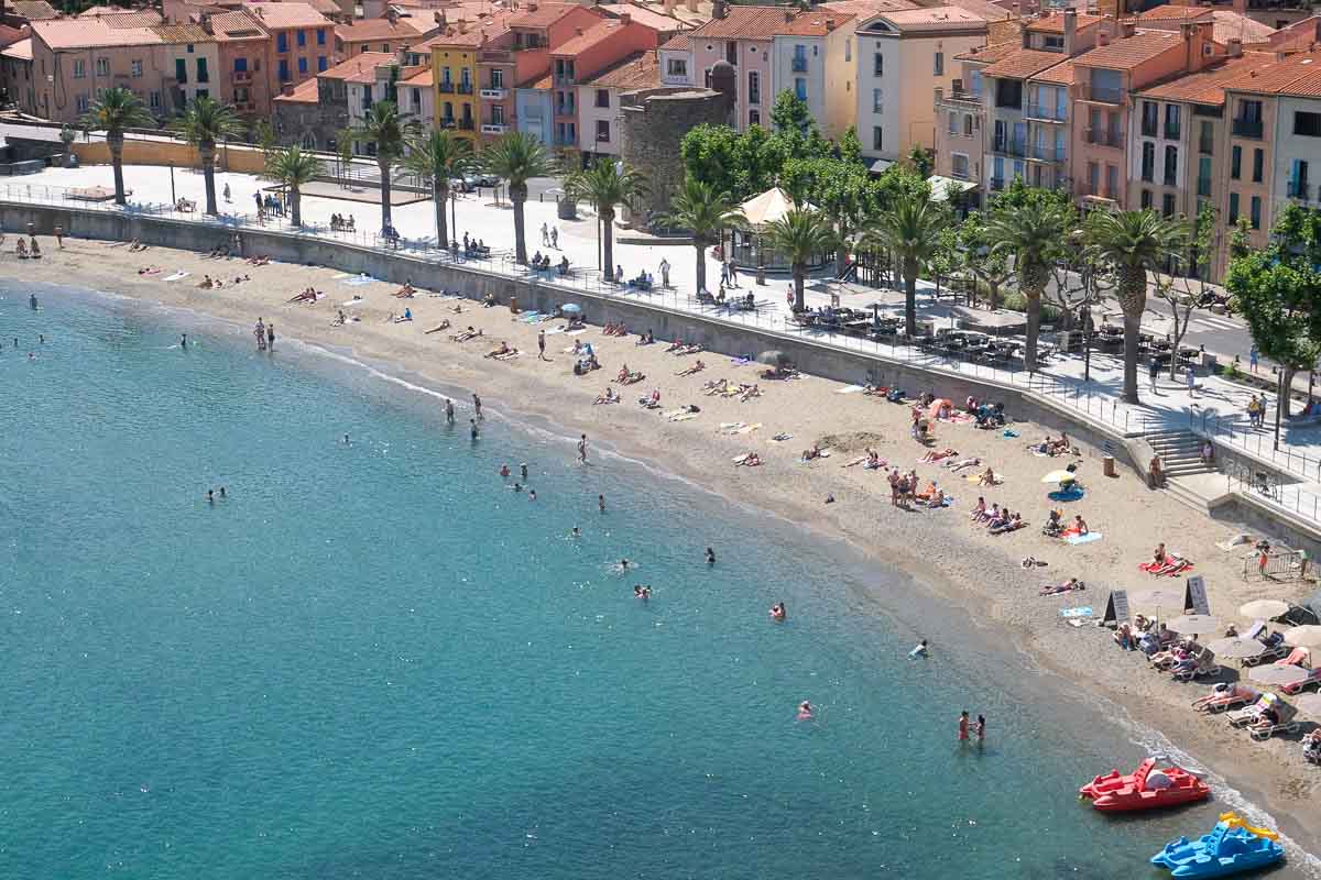 people sitting on beach and swimming in the sea backed by a promenade with pastel coloured buildings