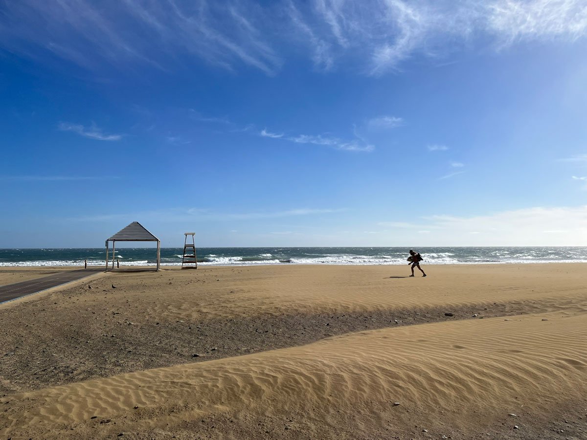 man walking across a broad sandy beach carrying a surf board