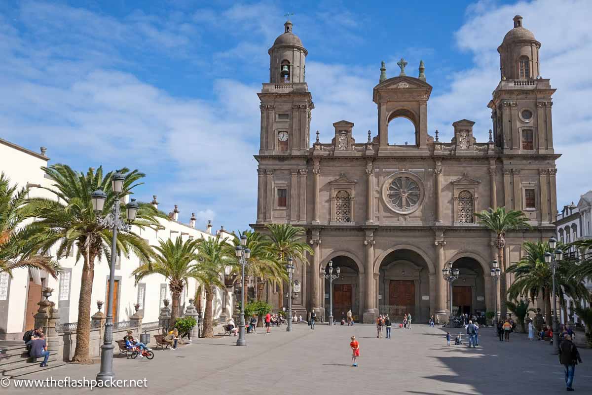 dark stone exterior of las palmas cathedral with 2 bell towers fronted by a large square with palm trees