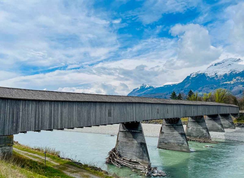 wooden covered bridge across an aquamarine river with snow covered mountains close by