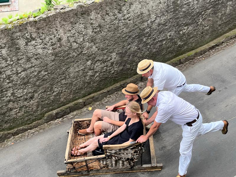 two men dresssed in white pushing a wicker toboggan at speed down a hill in madeira madeira