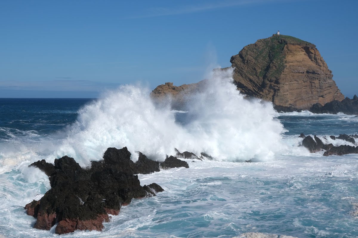 waves crashing against rocks with a small raised island with a lighthouse in madeira portugal