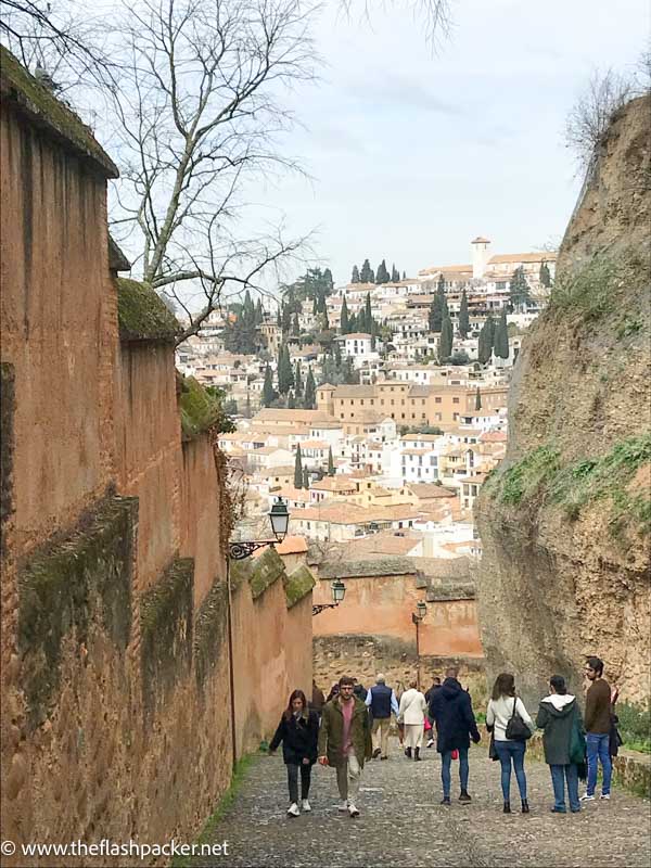 people walking along a steep path with stone walls on either side and the rooftops of granada in background