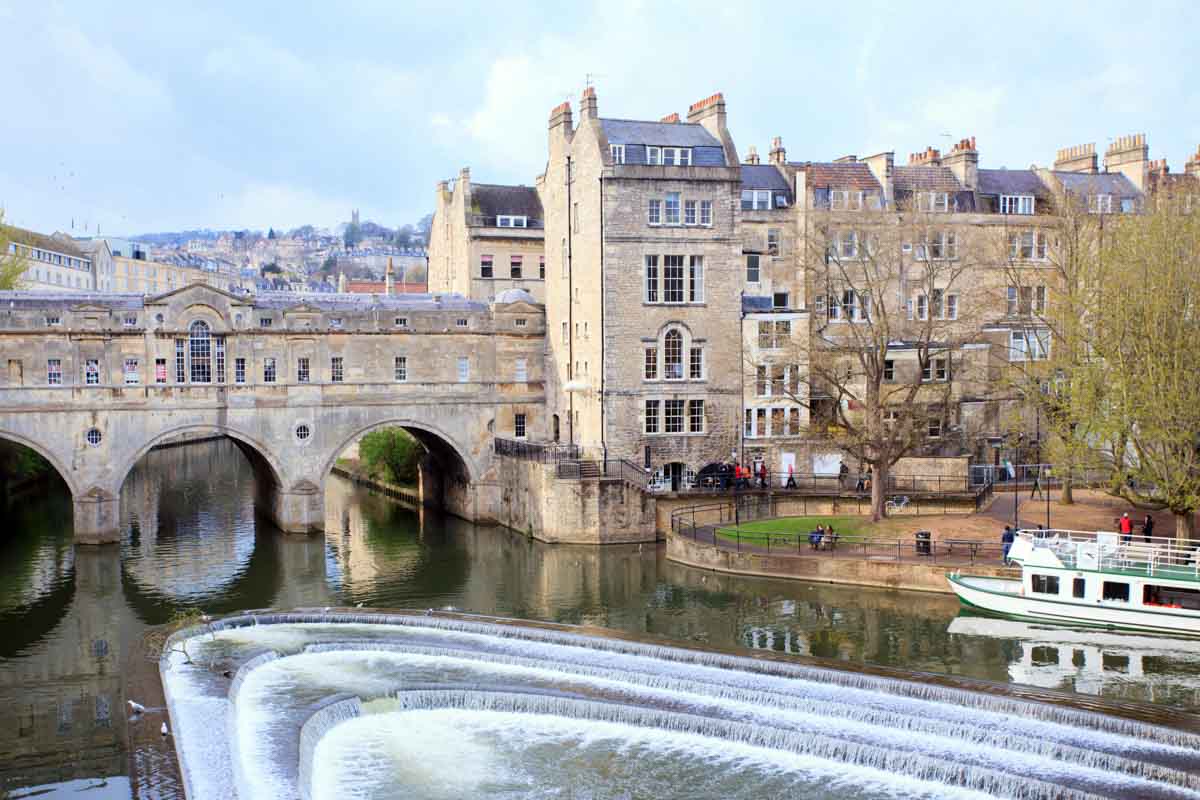 stone buildings and a covered bridge over a river with a weir and boat in bath england