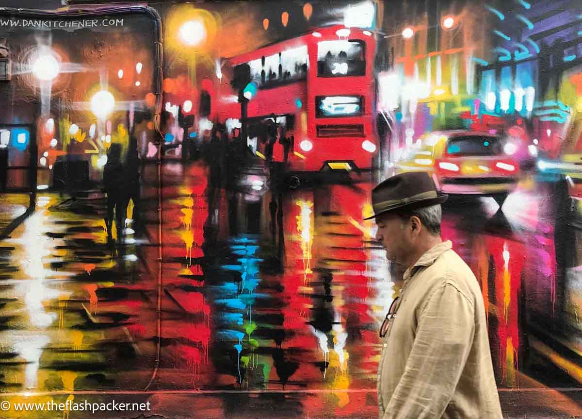 man walking by London scene painted on a wall of a red bus and cars reflected in a city street at night