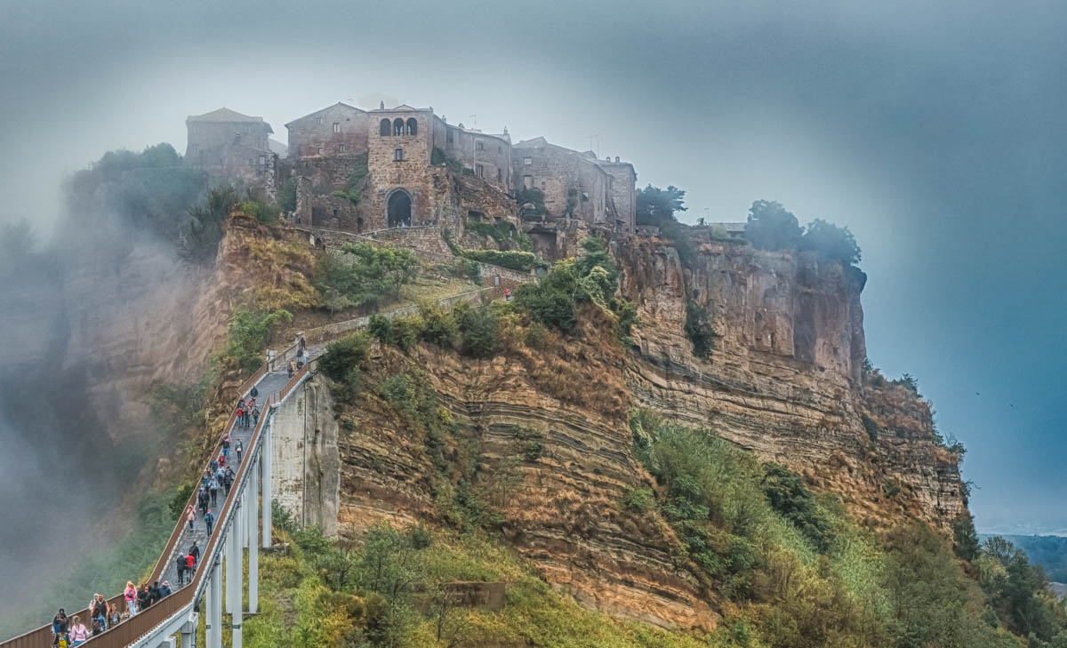 people walking along a footbridge to the old town of civita di bagnoregio on the top of a hill