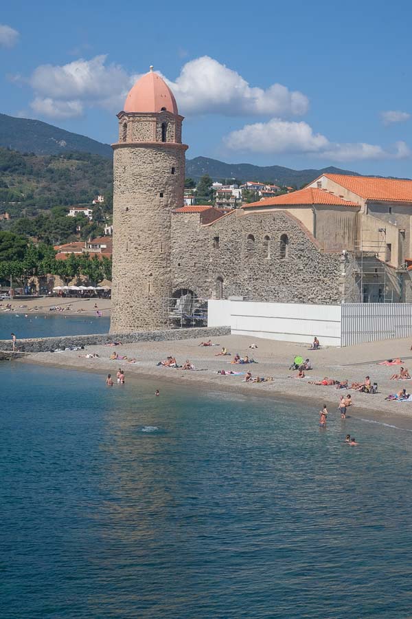 people by a small beach next to a a stone church with round bell tower