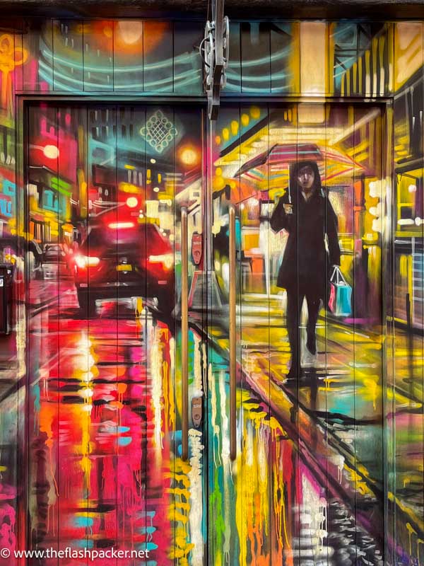 vibrant painting of person walking along a city street at night in rain carrying an umbrella