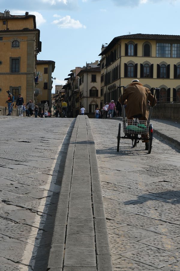 man pedalling a bicycle and cart across a stone foot bridge in florence