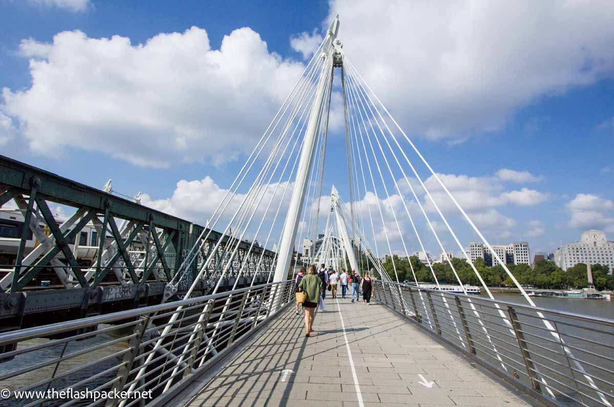 people walking across suspension bridge in london with white cables on a sunny day