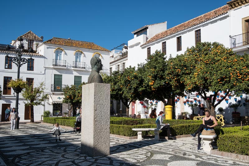 people in pretty square with a central statue of mans head and orange trees