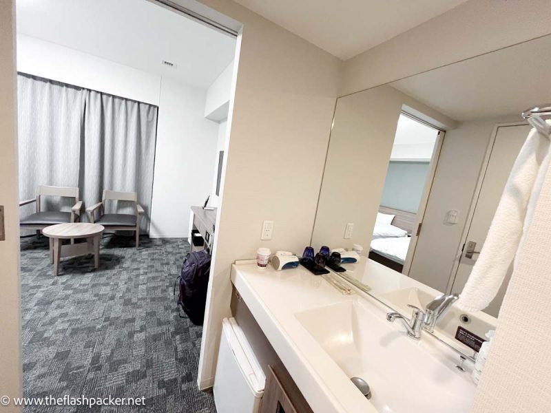 white hotel bathroom looking into bedroom with grey furnishings