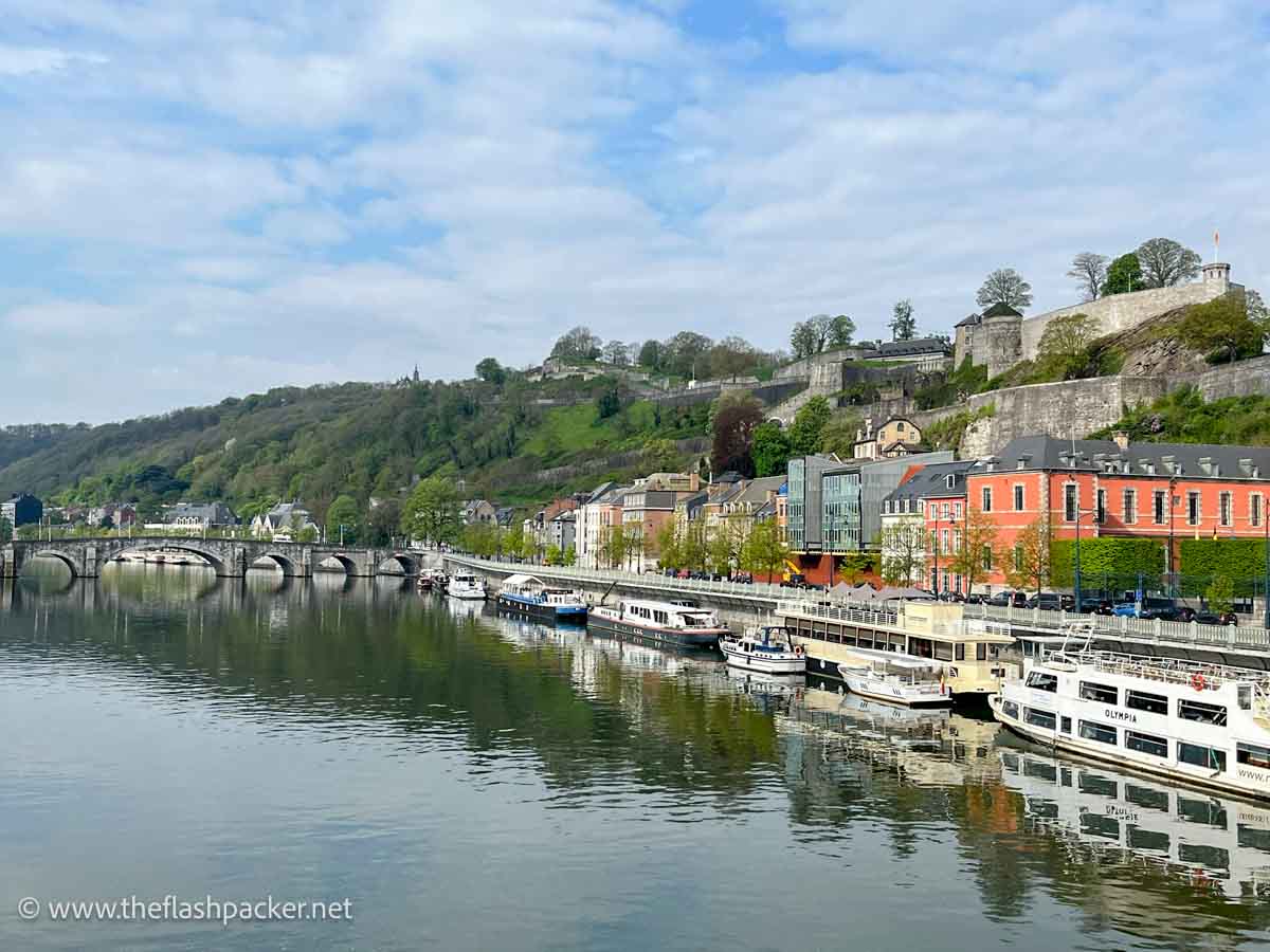 river in namur lined with pleasure boats and vibrant buildings and a castle on the hill