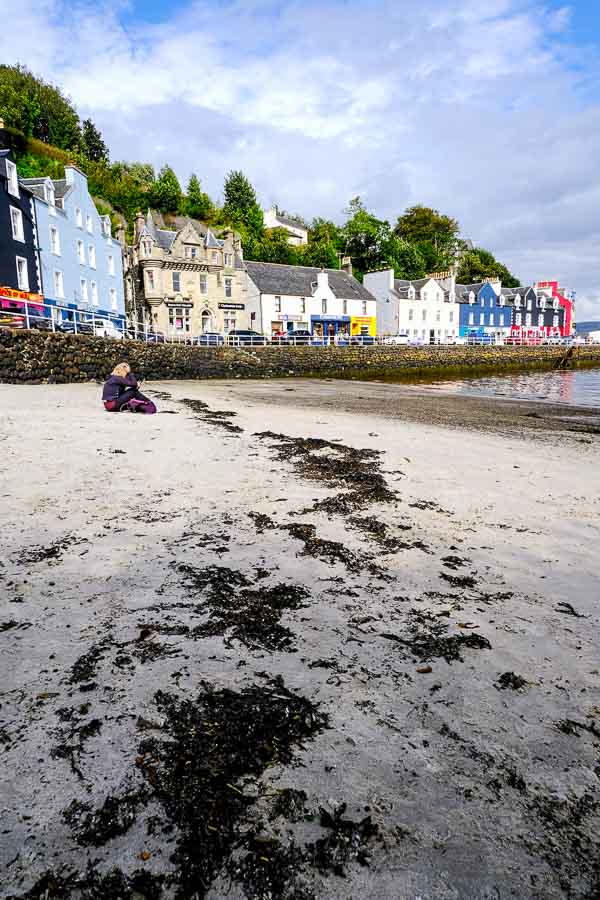 woman sitting on sand in tobermory with brightly coloured houses in background