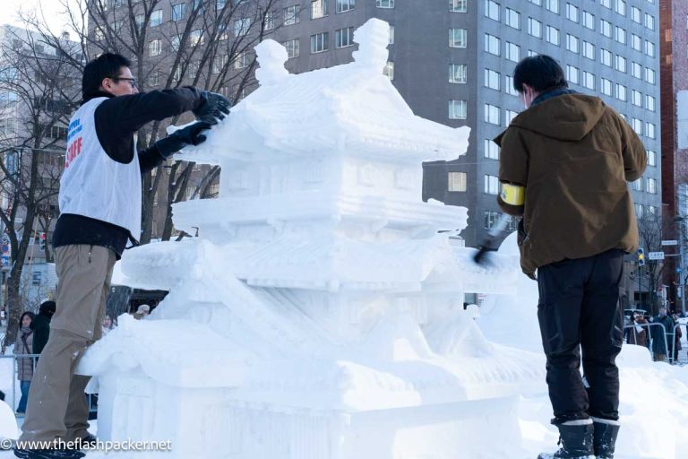 2 men carving snow around a sculpture of a castle