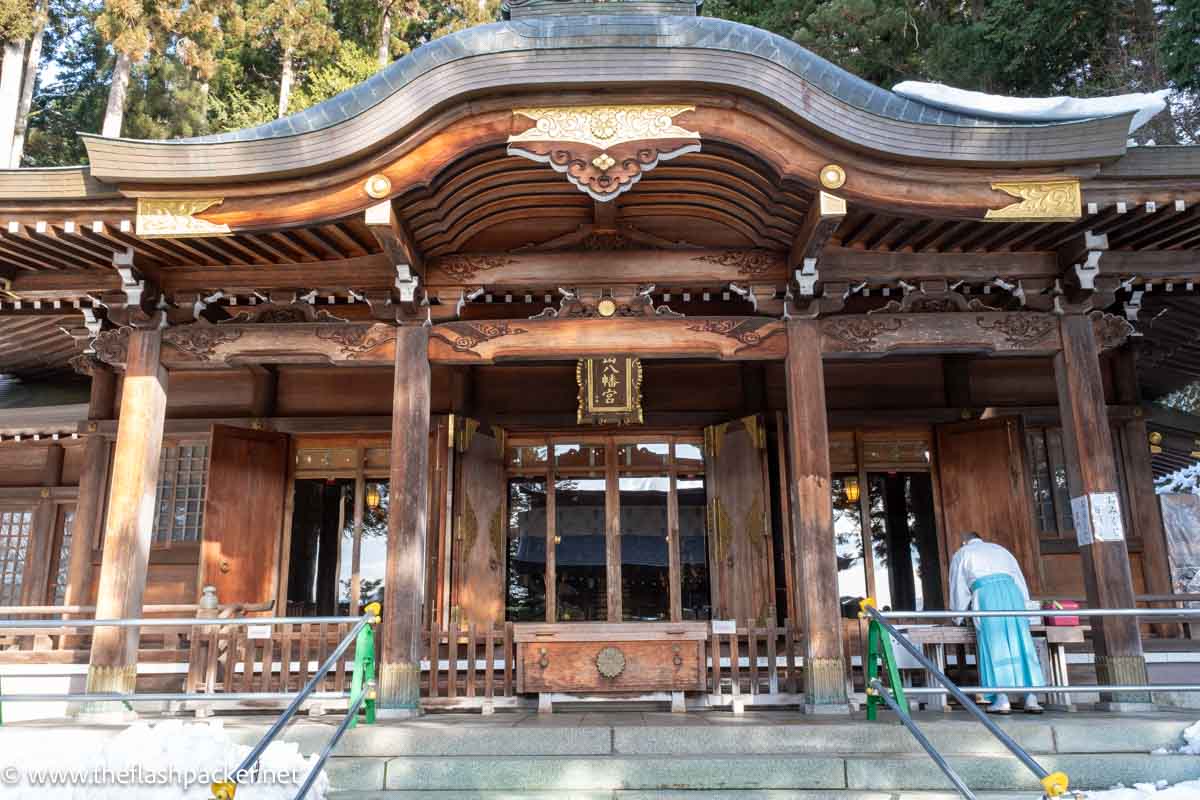 man in blue and white robes outside a wooden shinto shrine