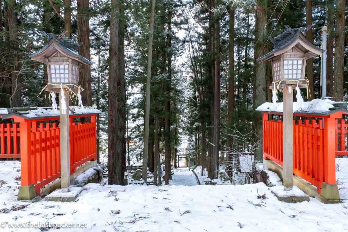 2 lamp posts and red painted gates at the entrance to a snowy forest path