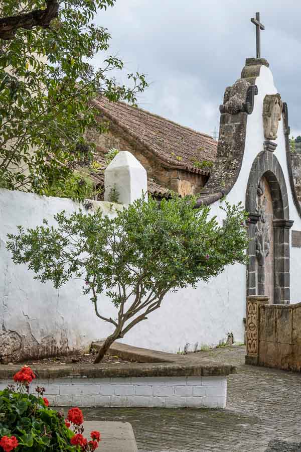 stone arch and gateway with a cross on top and a small plaza with red flowers and little tree