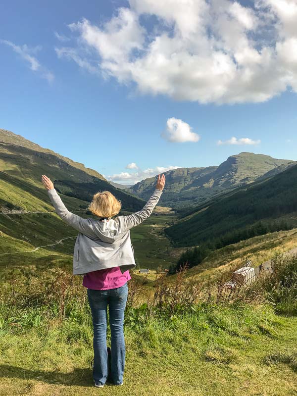 a woman who is a solo traveller in her 50s standing with arms outstretched in a scottish valley