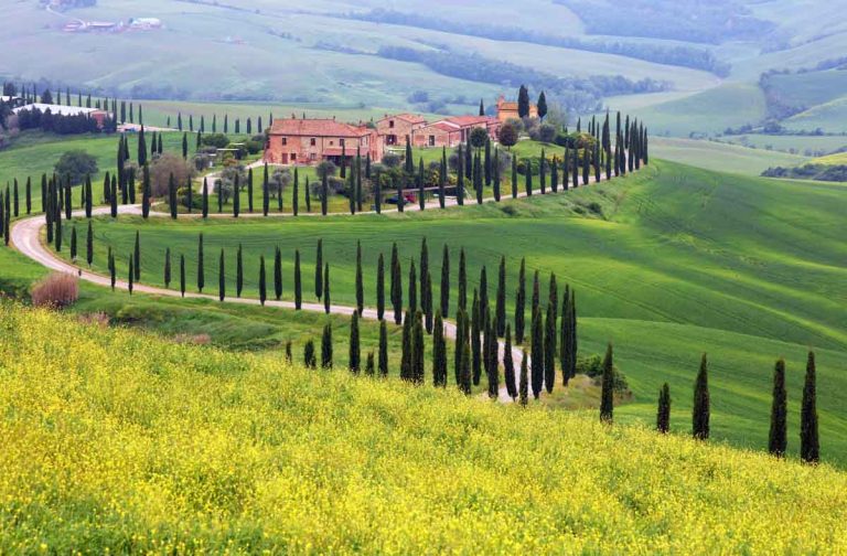 rolling landscape of tuscany italy with winding path lined with cypress trees leading to red-brick houses