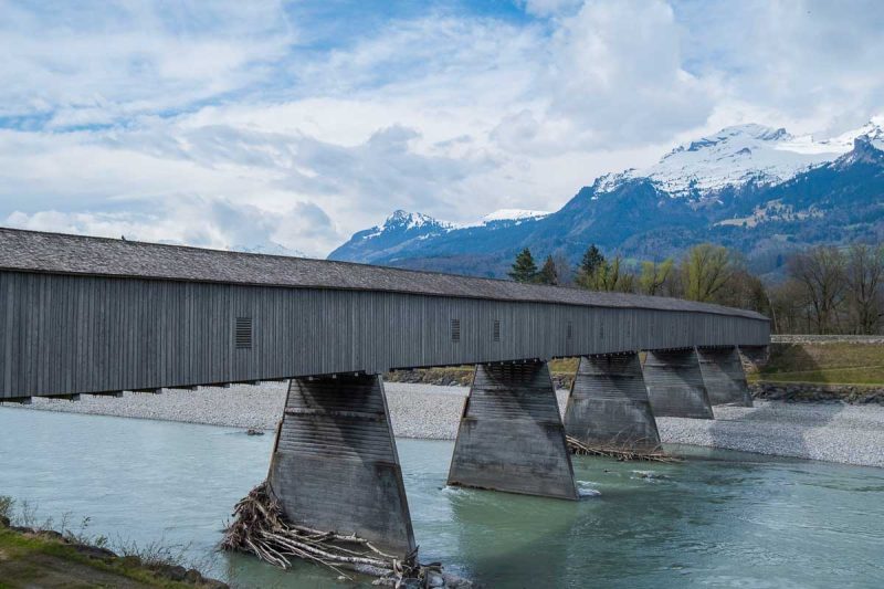 wooden covered bridge across an aquamarine river with snow covered mountains close by