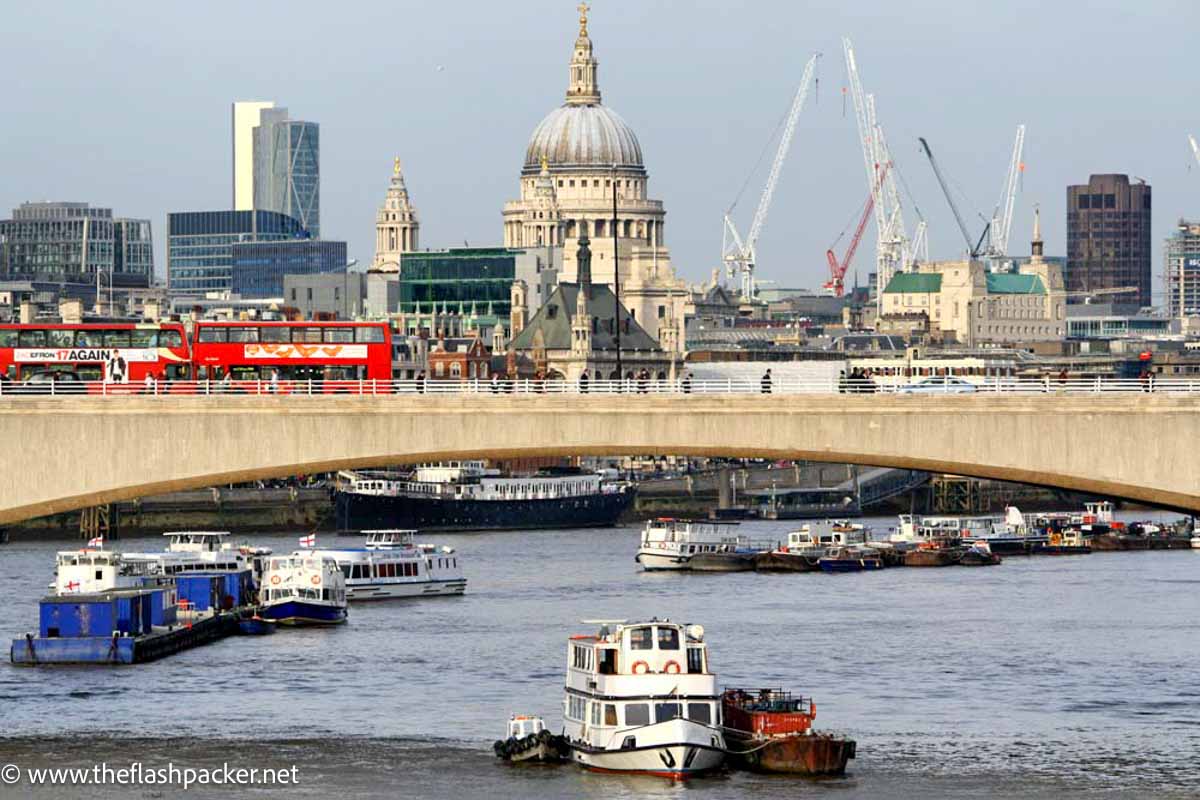 concrete bridge over river thames in london with red buses and cathedral dome in background