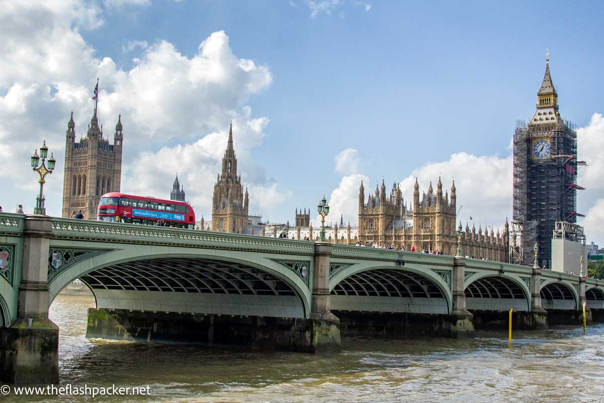 red bus crossing bridge over the thames in london with houses of parliament and big ben